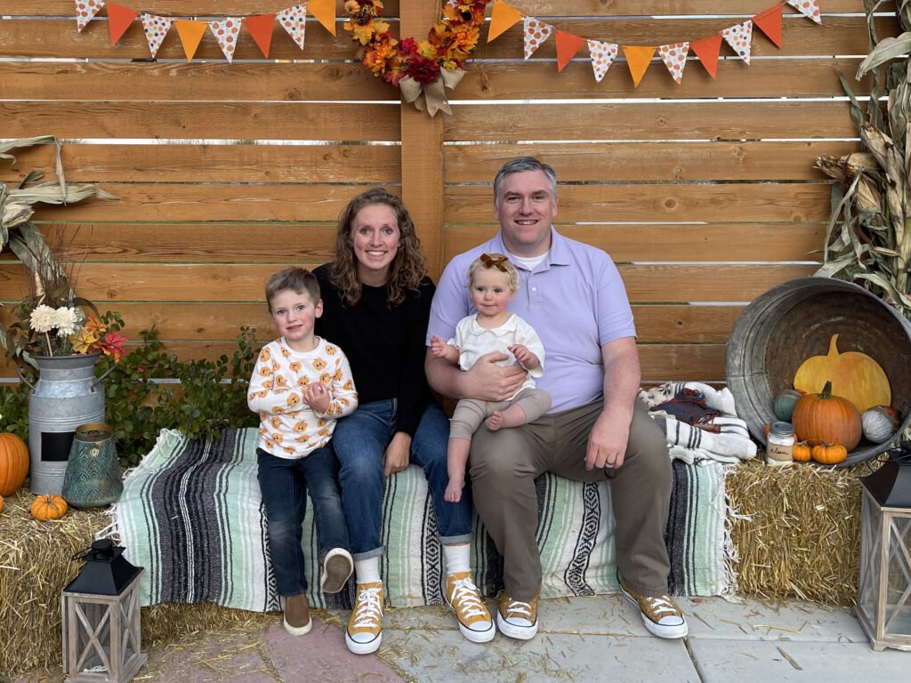 Harrison family sitting on a hay bale at a fall festival. Nathan, Mackenzie, and 2 children.